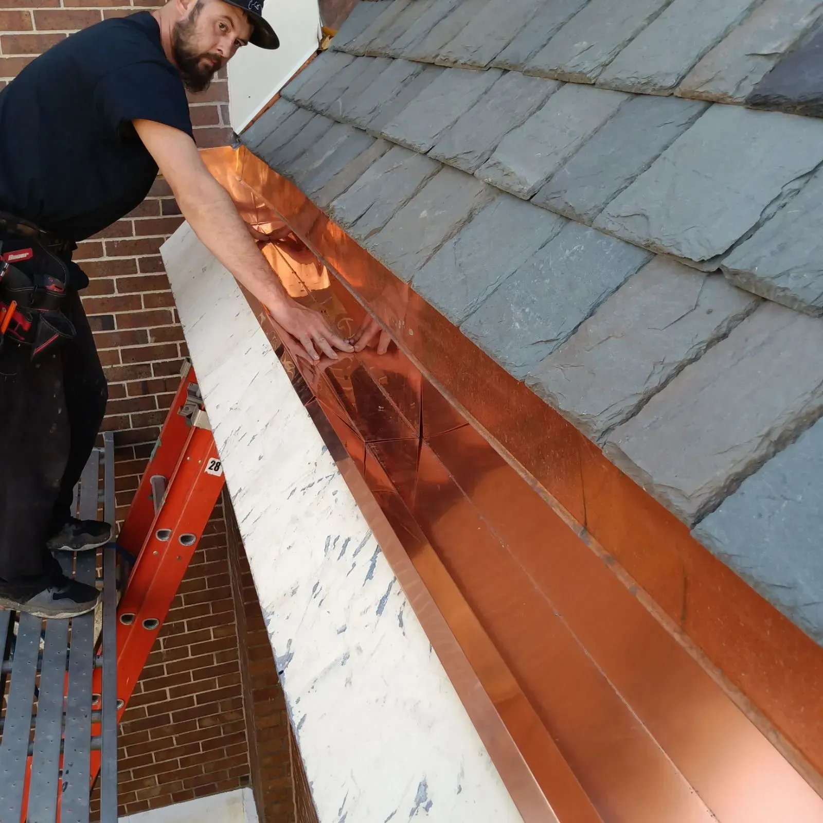 Worker installing copper flashing on a slate roof beside a ladder