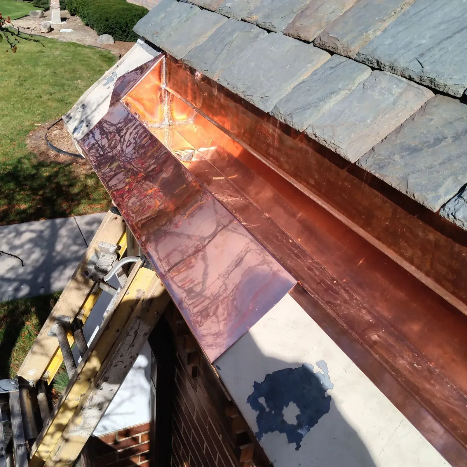 Copper roof flashing being installed along a slate roof edge next to a ladder.