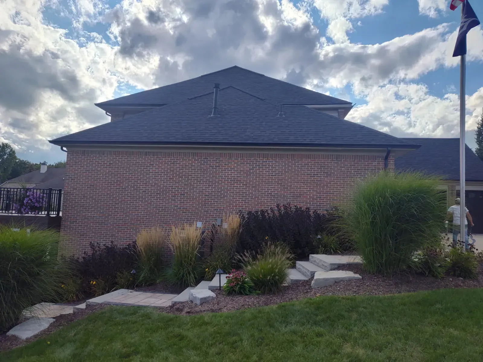 Brick building with dark shingled roof, landscaped garden, and cloudy sky.