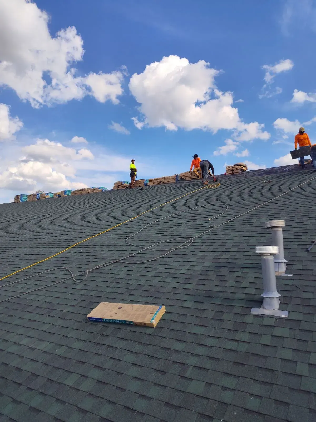 Roofing crew working on a steep shingled roof under a blue sky with clouds.