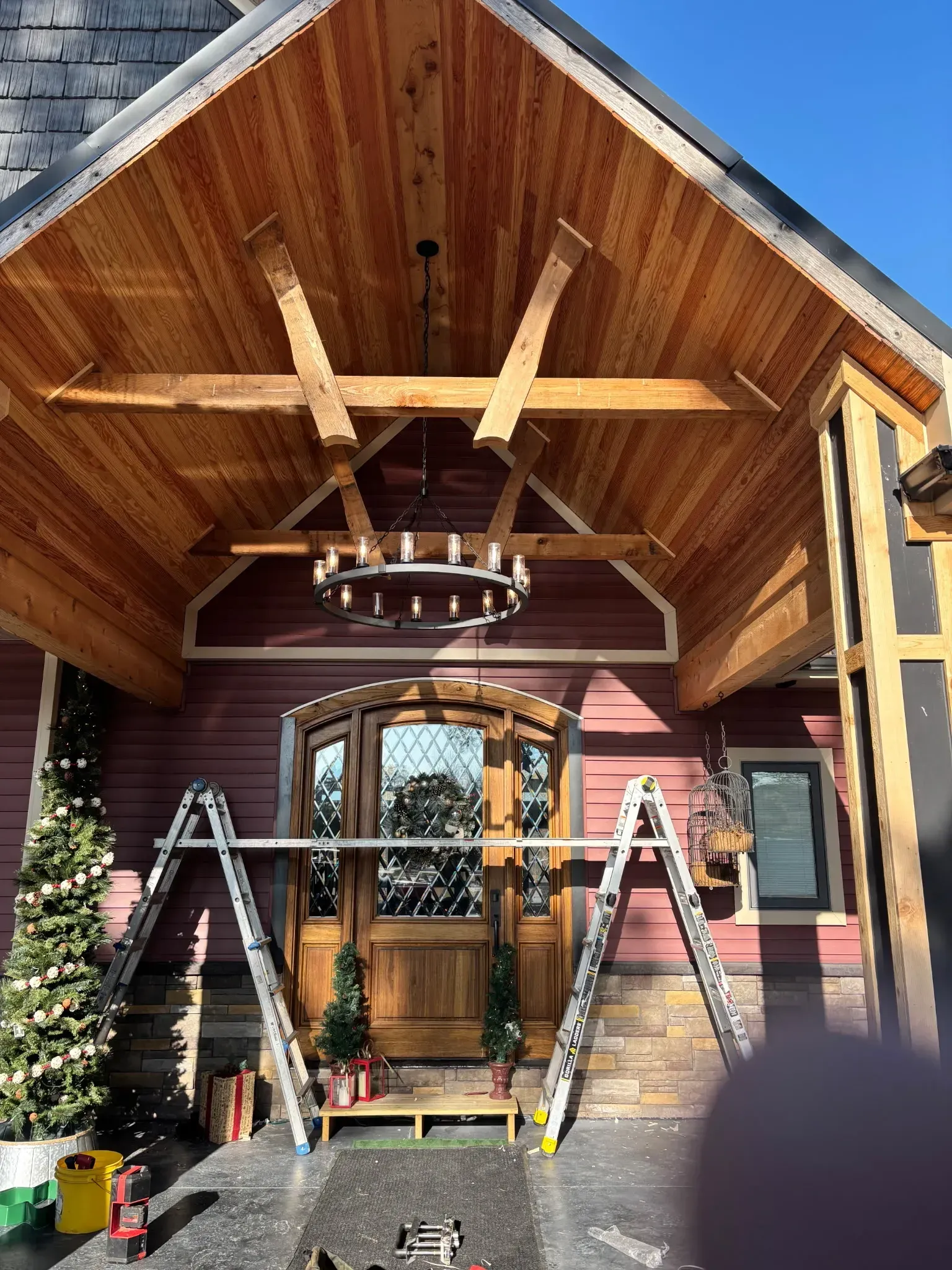 Front porch with steep wood gable, chandelier, double doors, and ladders set up under construction.