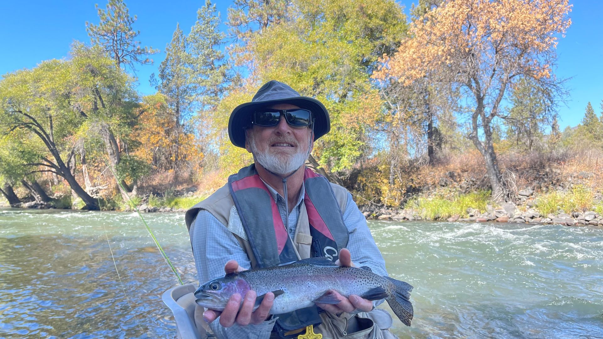 Man holding rainbow trout, wearing hat, vest, sunglasses, smiling on a river, autumn foliage.