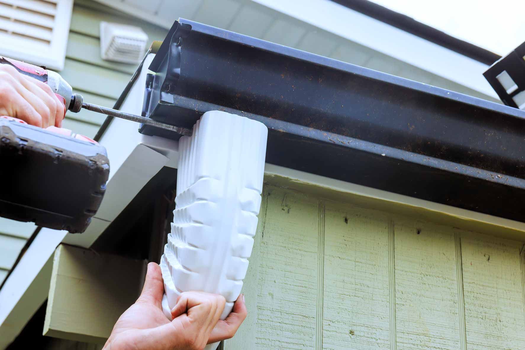 Person installing a white downspout under a roof gutter on a house exterior