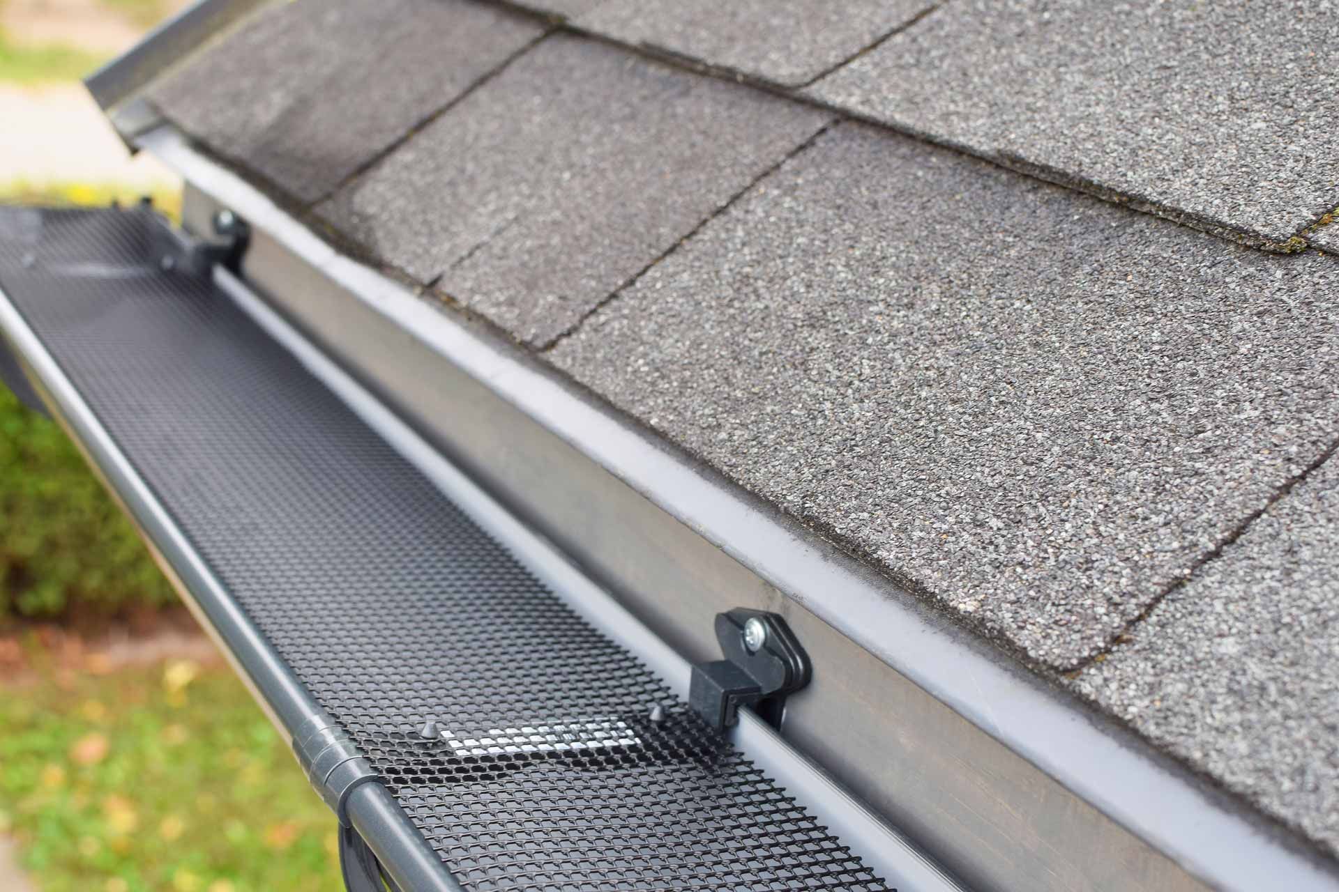 Gutter guard installed along a shingled roof edge, with downspout bracket and mesh debris screen.