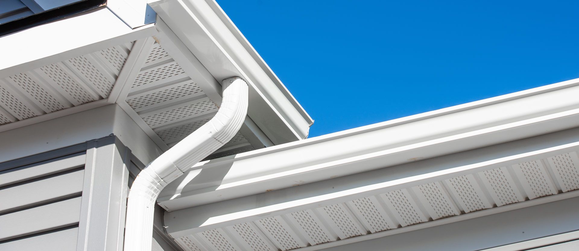 White rain gutters and downspout on a gray house roof against a blue sky