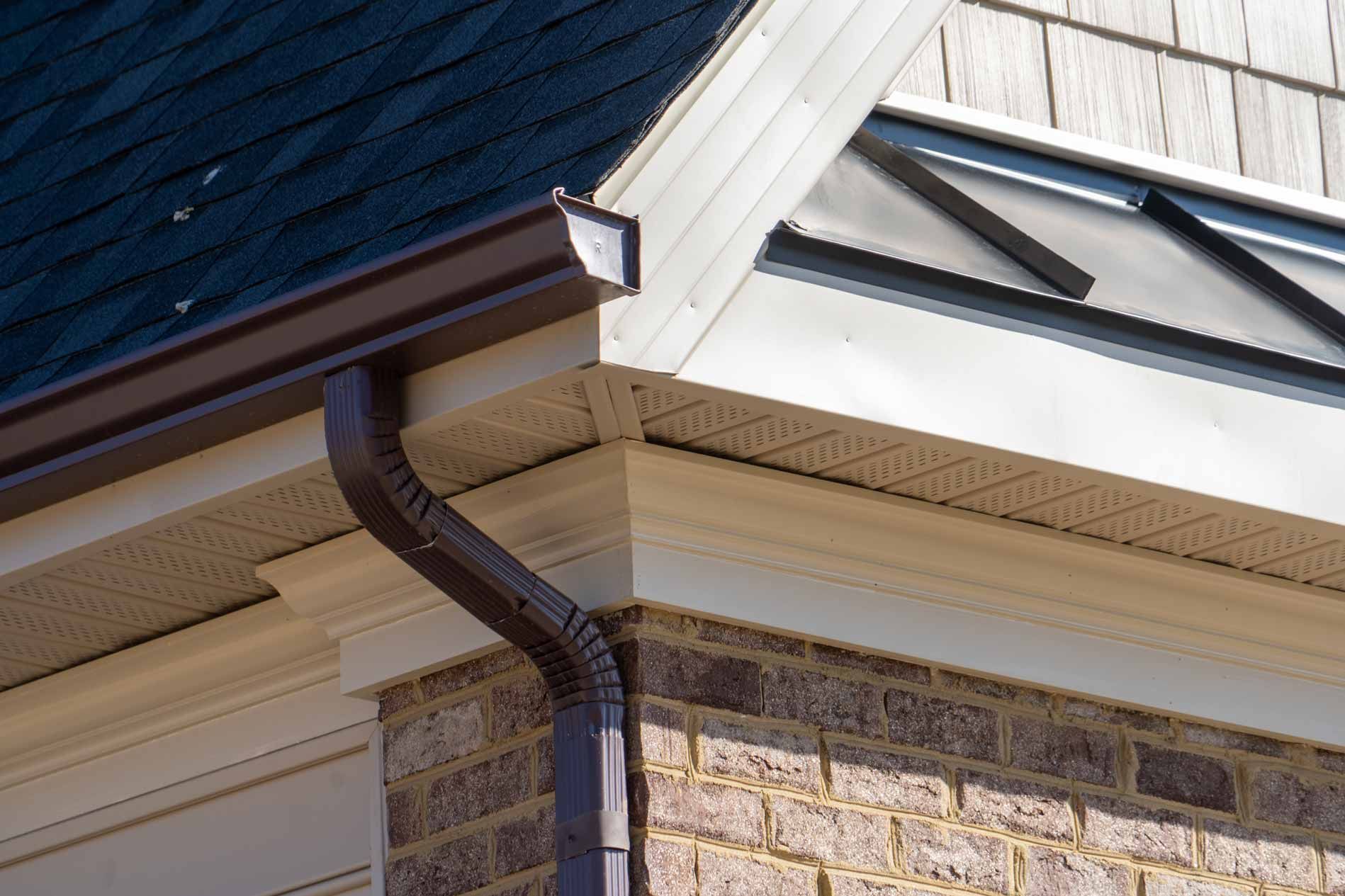 Roof corner with beige trim, dark shingle siding, and a brown gutter downspout on a stone wall