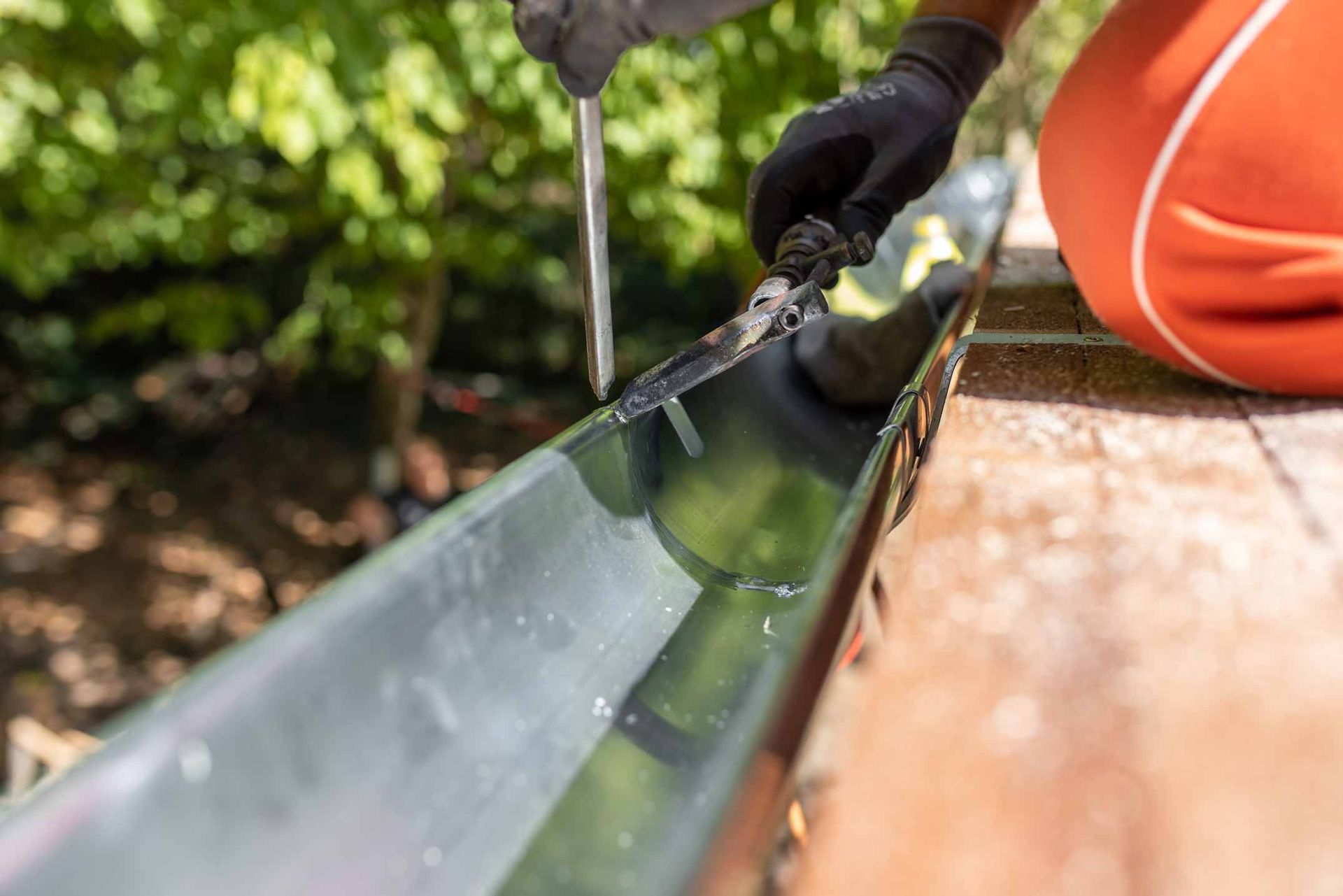 Gloved hands cleaning a rain gutter beside an orange bucket outdoors