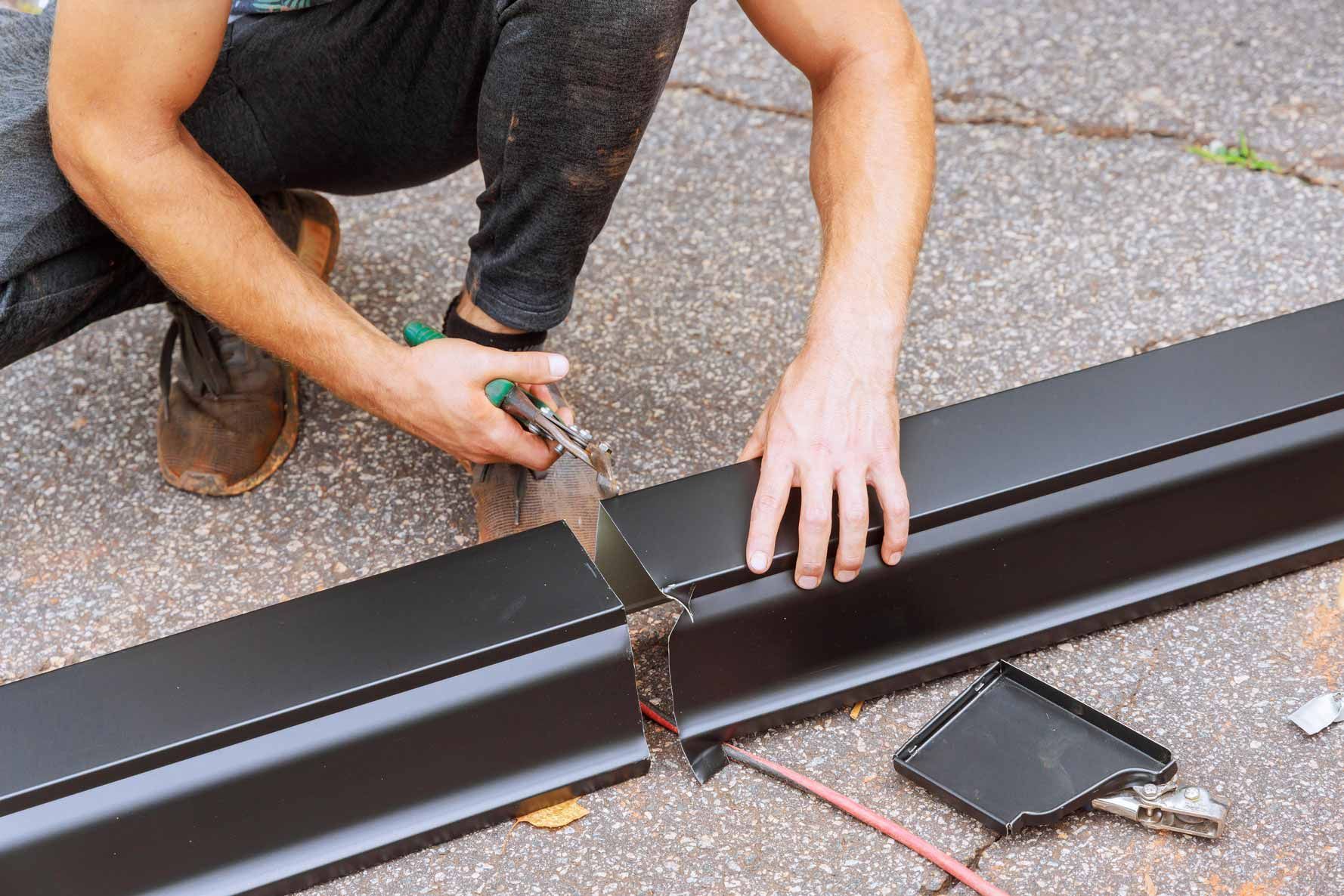 Person cutting a black metal gutter section with a saw on a driveway
