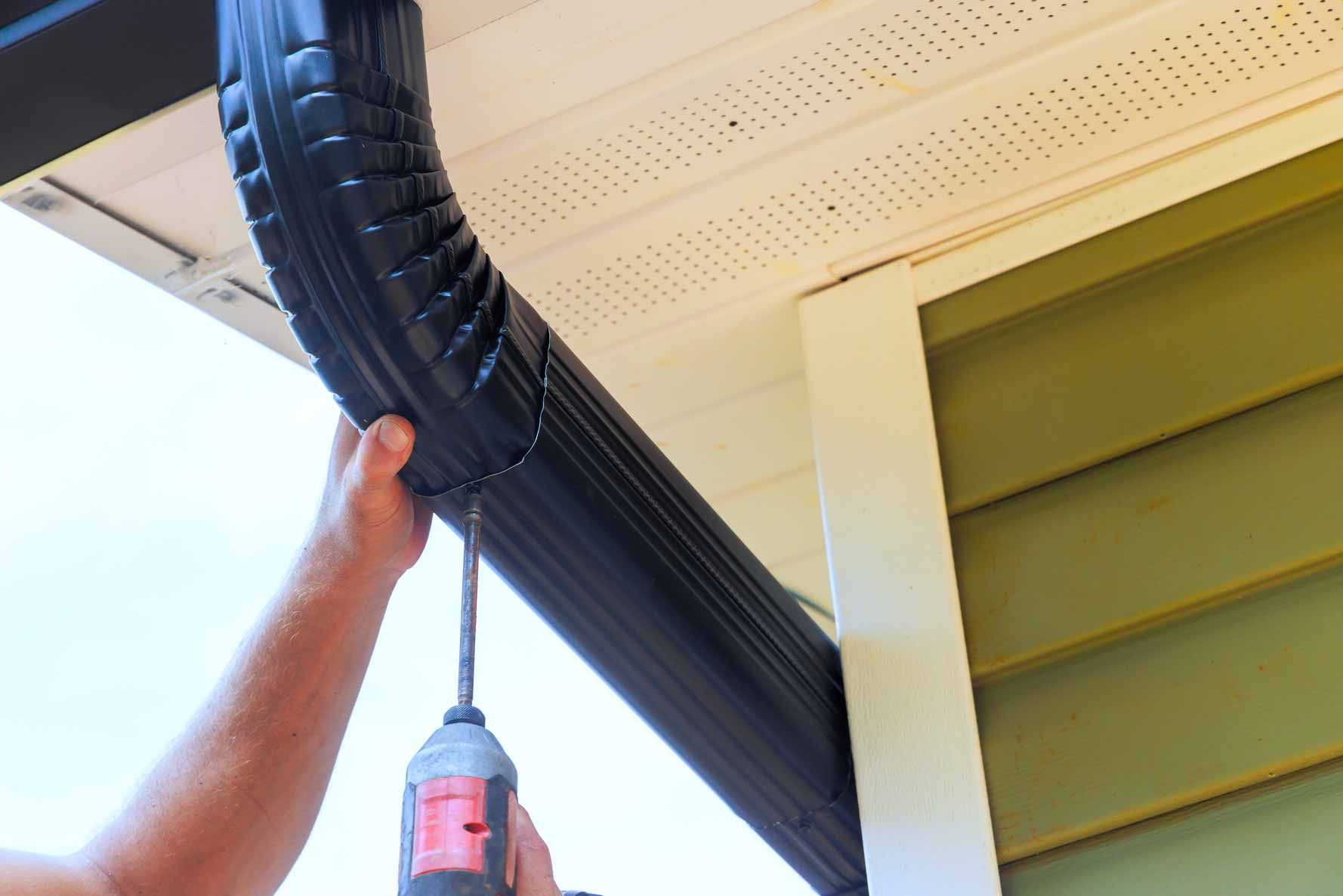Hand holding black downspout gutter under a house eave, using a cordless drill to fasten it