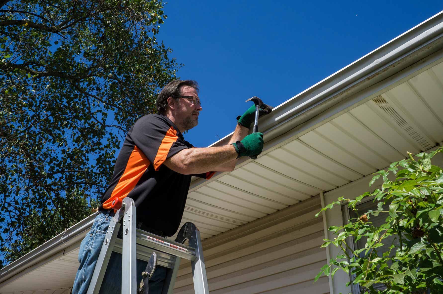 Man on a ladder cleaning gutters on a house roofline under a clear blue sky