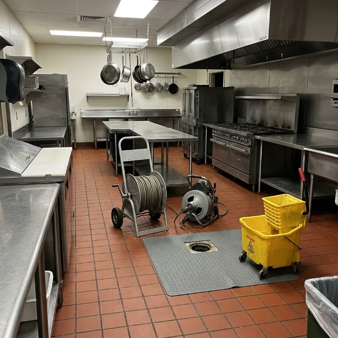 Industrial kitchen with stainless steel counters, hanging pots, prep tables, and yellow cleaning cart on tiled floor