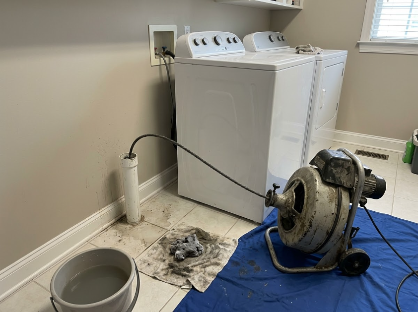 Laundry room with a washing machine, clogged drain, wet floor, and a drain-cleaning auger on a blue tarp