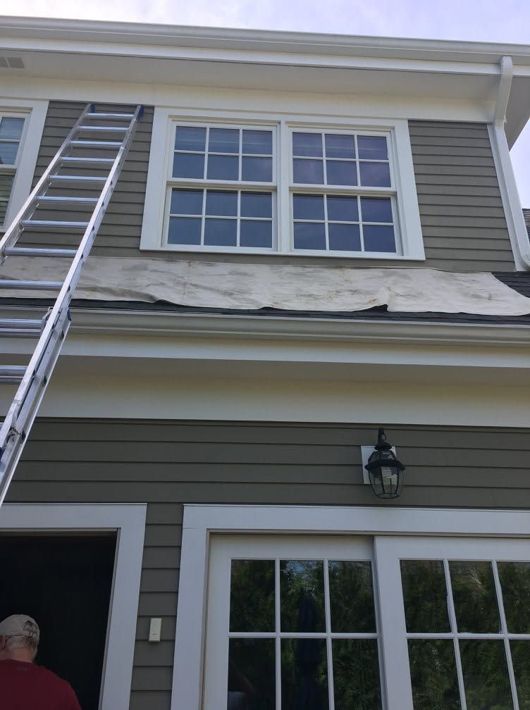 Exterior of a gray house with second-story windows, ladder, and damaged roof shingles above a porch