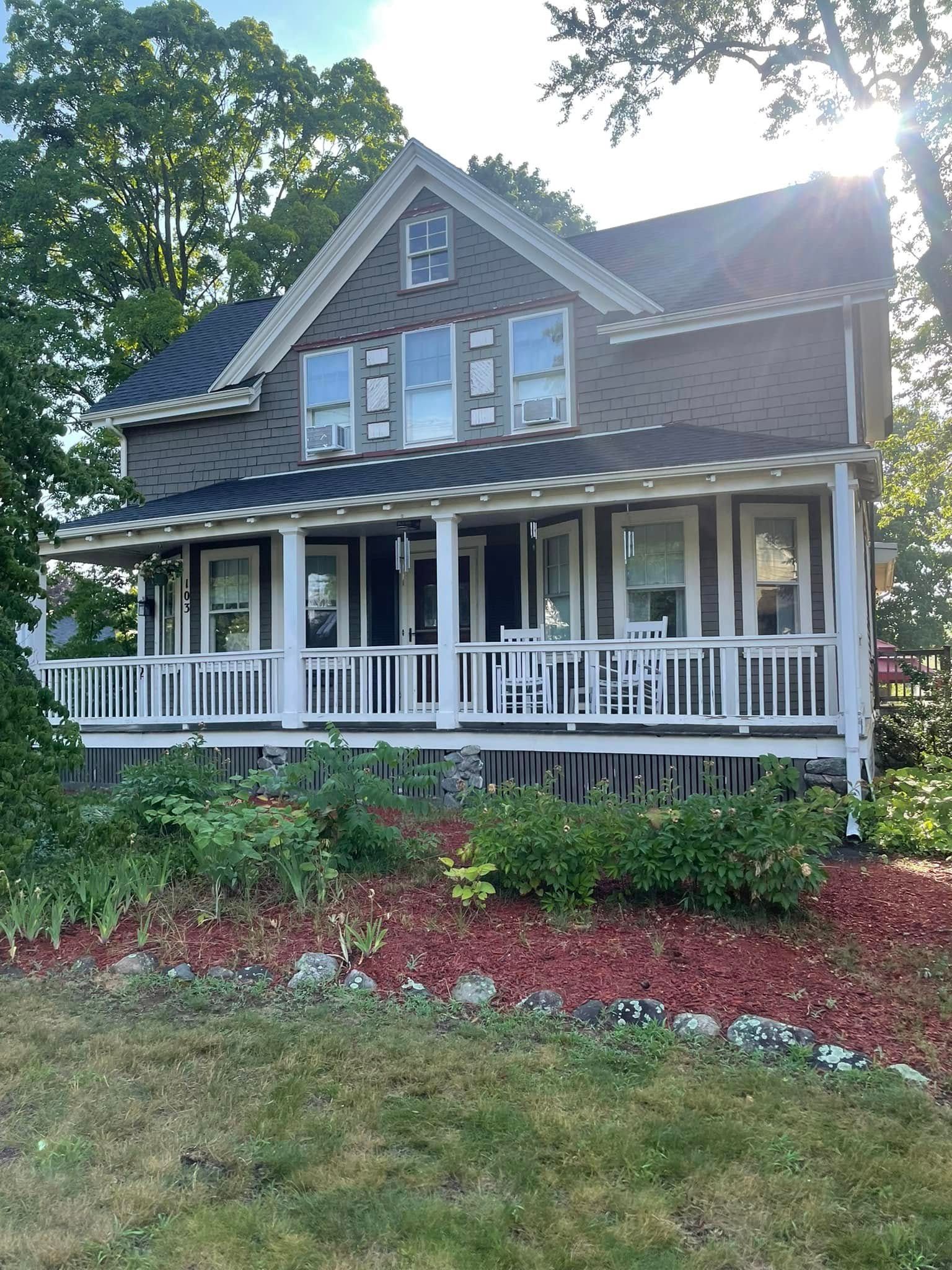 Victorian house with white porch railing, dark shingles, and a front yard garden in sunlight