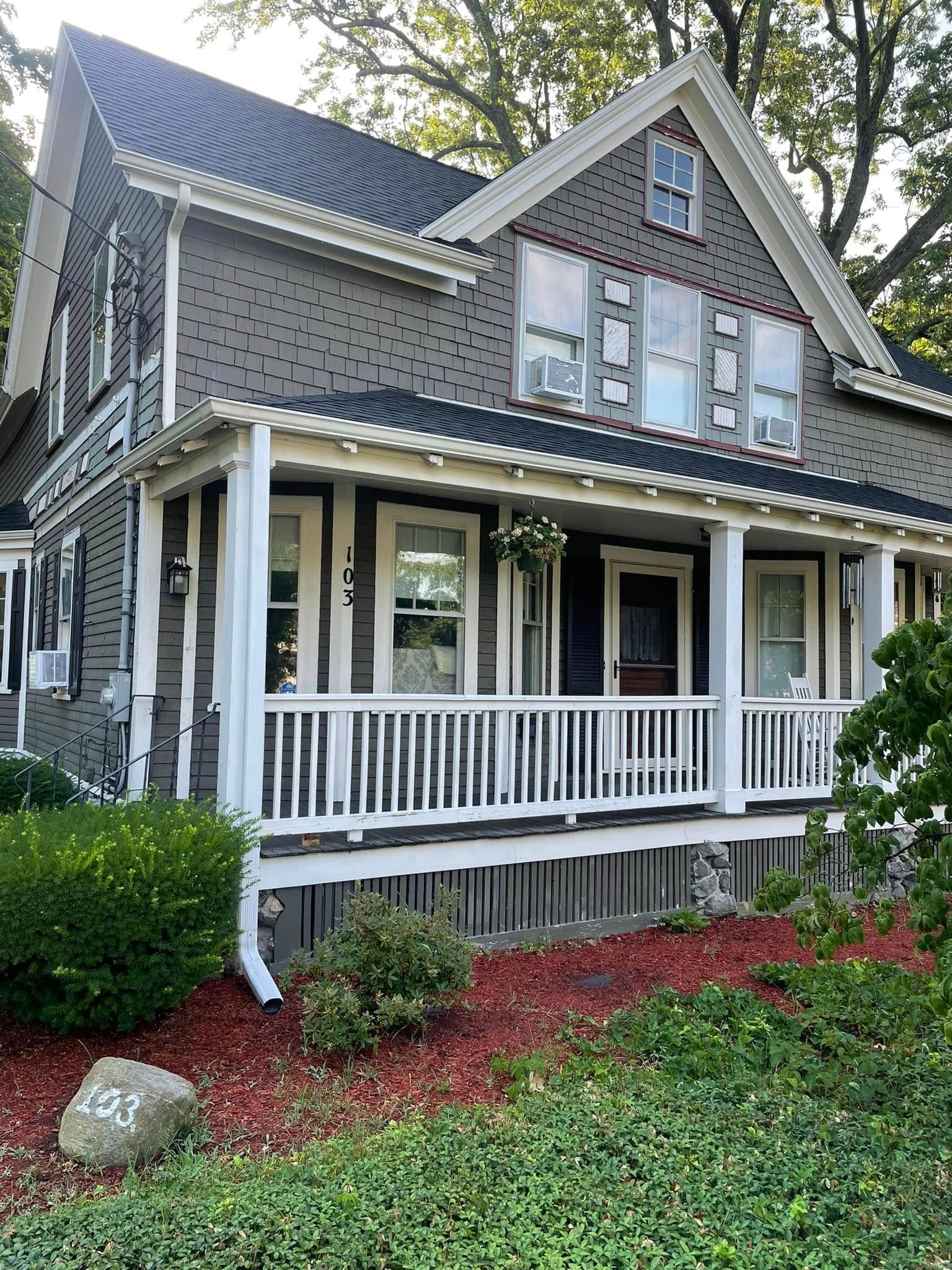 Two-story gray house with white porch railings, gabled roof, and landscaped front yard with mulch and shrubs
