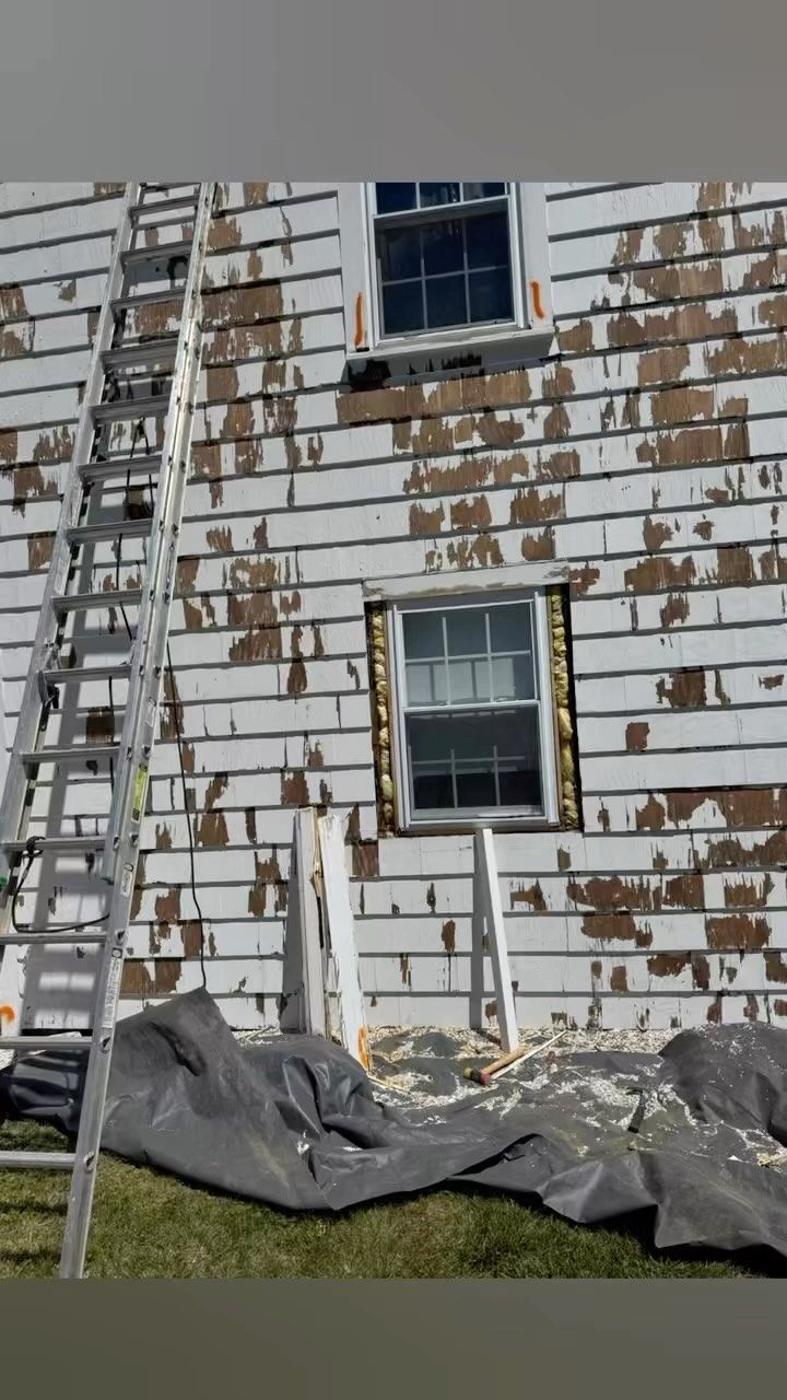 Weathered white house siding with peeling paint, two windows, and a tarp-covered area at the base