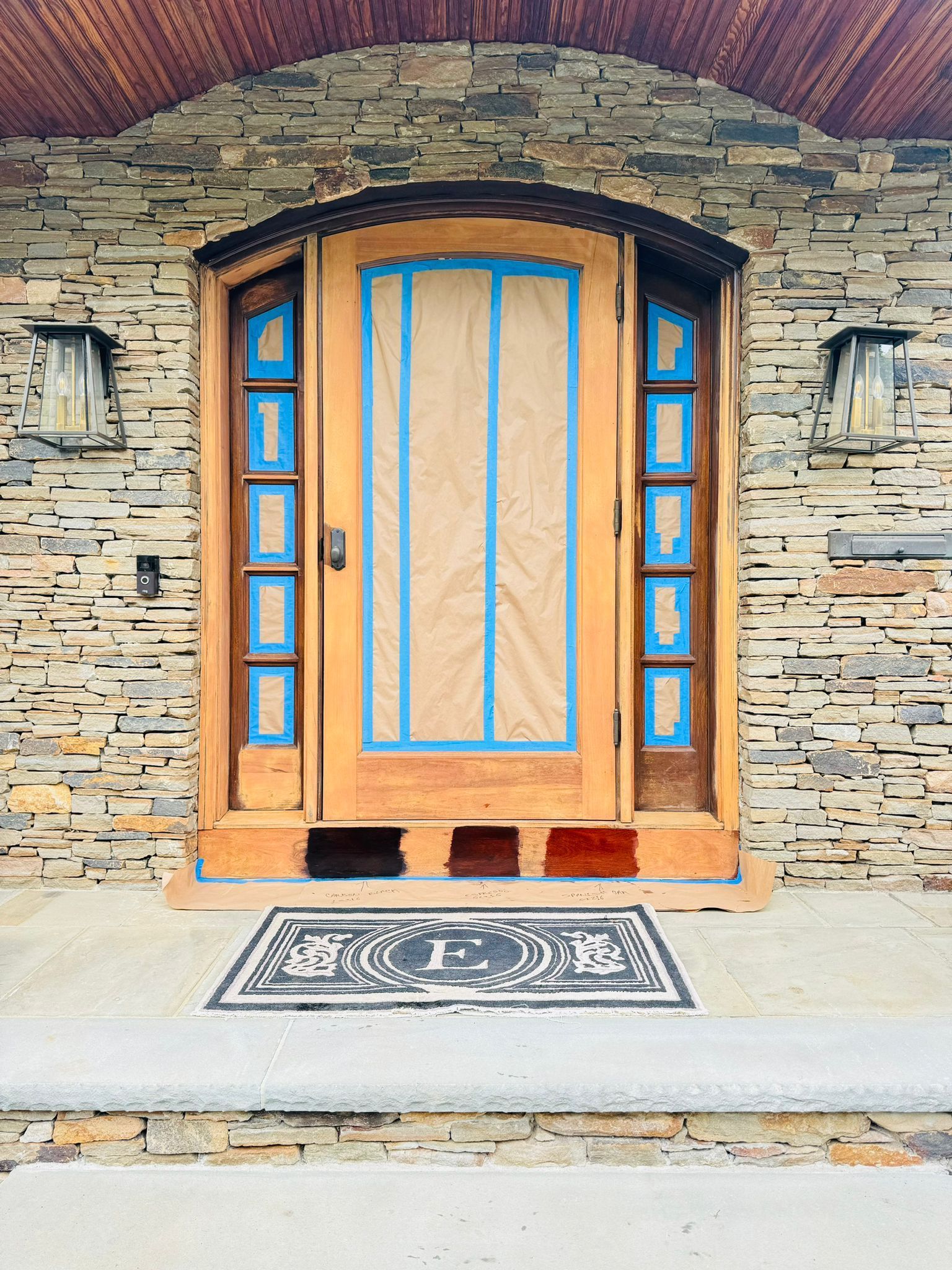 Wooden front door with blue trim set in a stone wall, with lanterns and a patterned doormat.