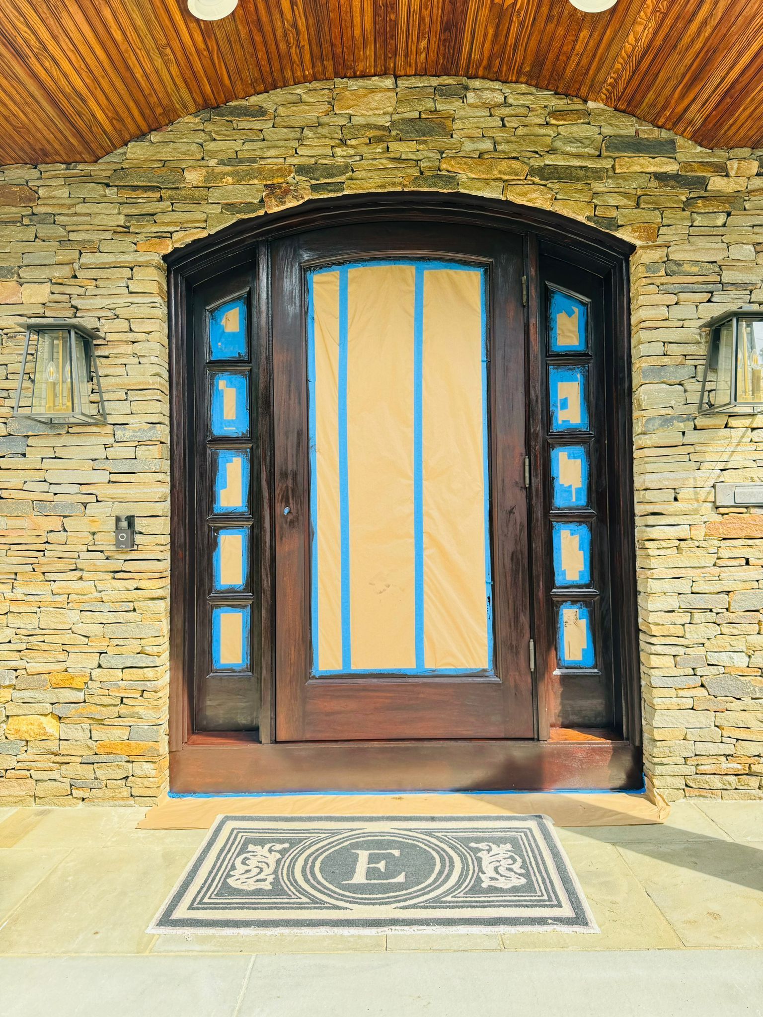 Stone entryway with arched wooden door, blue glass side panels, and a decorative doormat under a brick ceiling