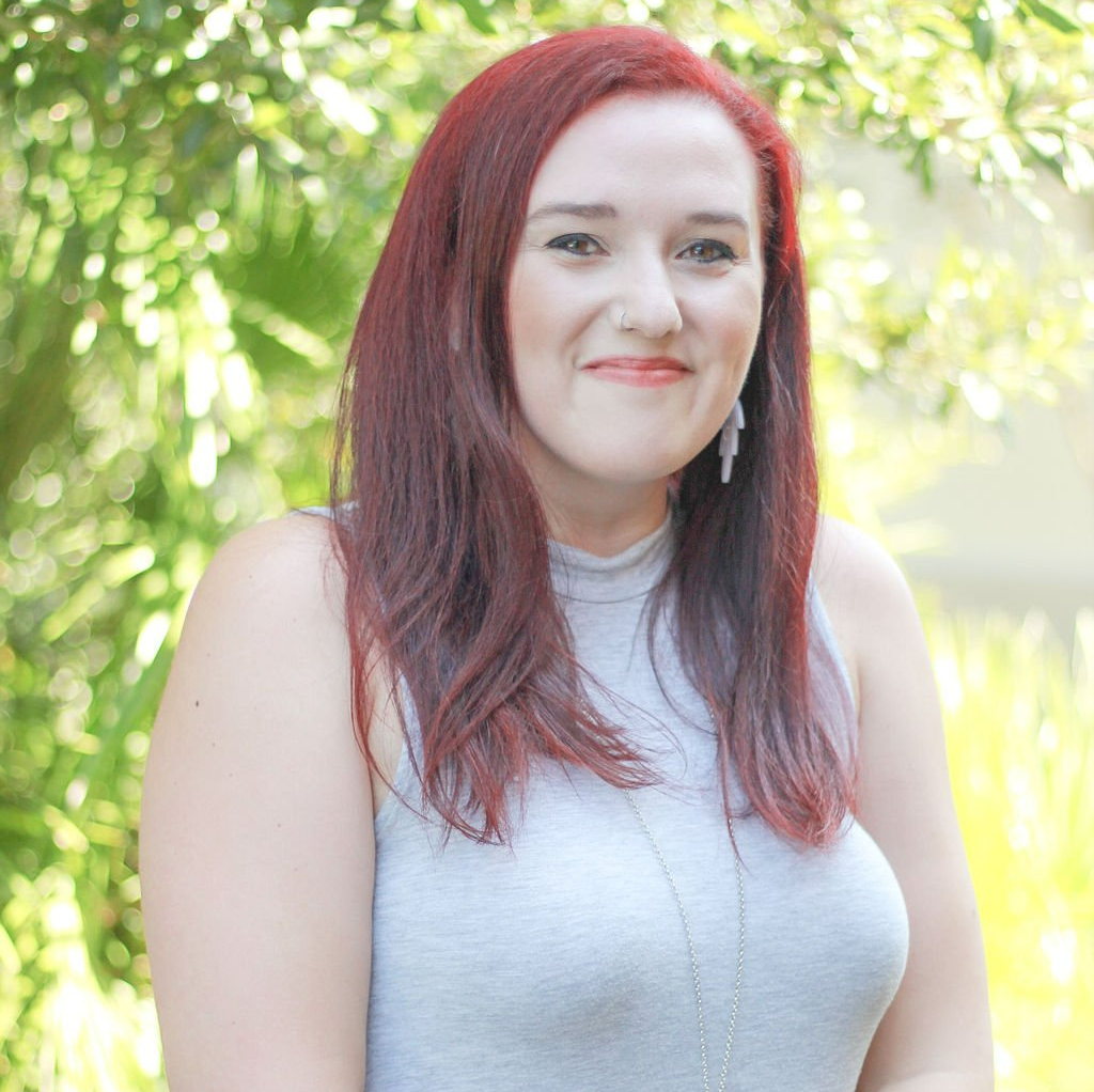 Woman with red hair wearing a gray top smiles in front of blurred greenery.