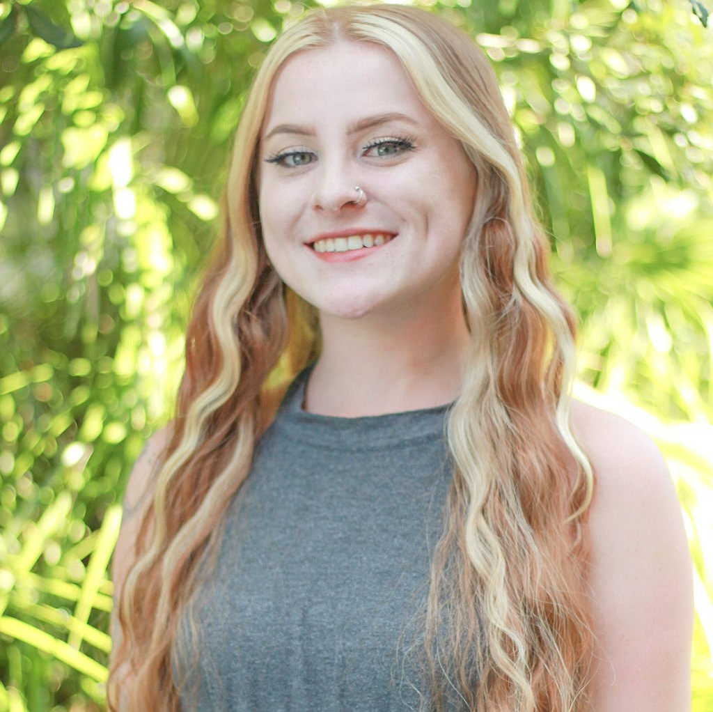 Young woman with blonde and brown hair smiles outdoors; wears a gray sleeveless top.