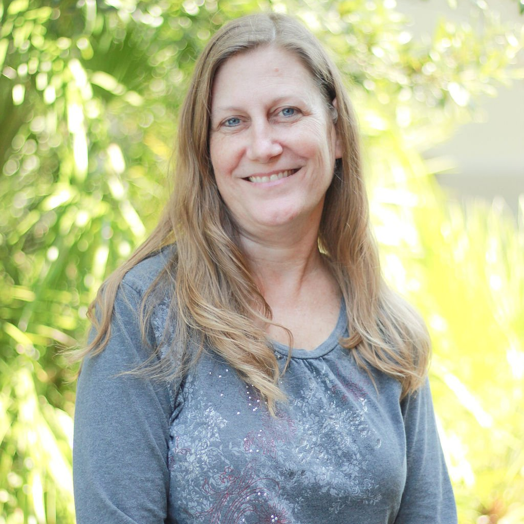Woman with long blond hair, smiling, wearing a gray shirt, standing outdoors in front of green foliage.