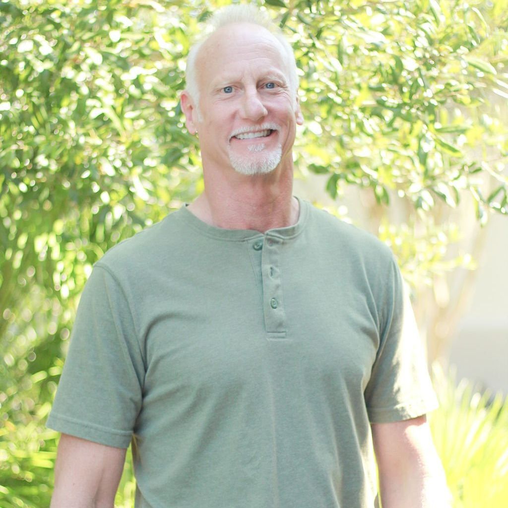 A smiling, middle-aged man with white hair and a goatee wears a green shirt, standing in front of a leafy background.