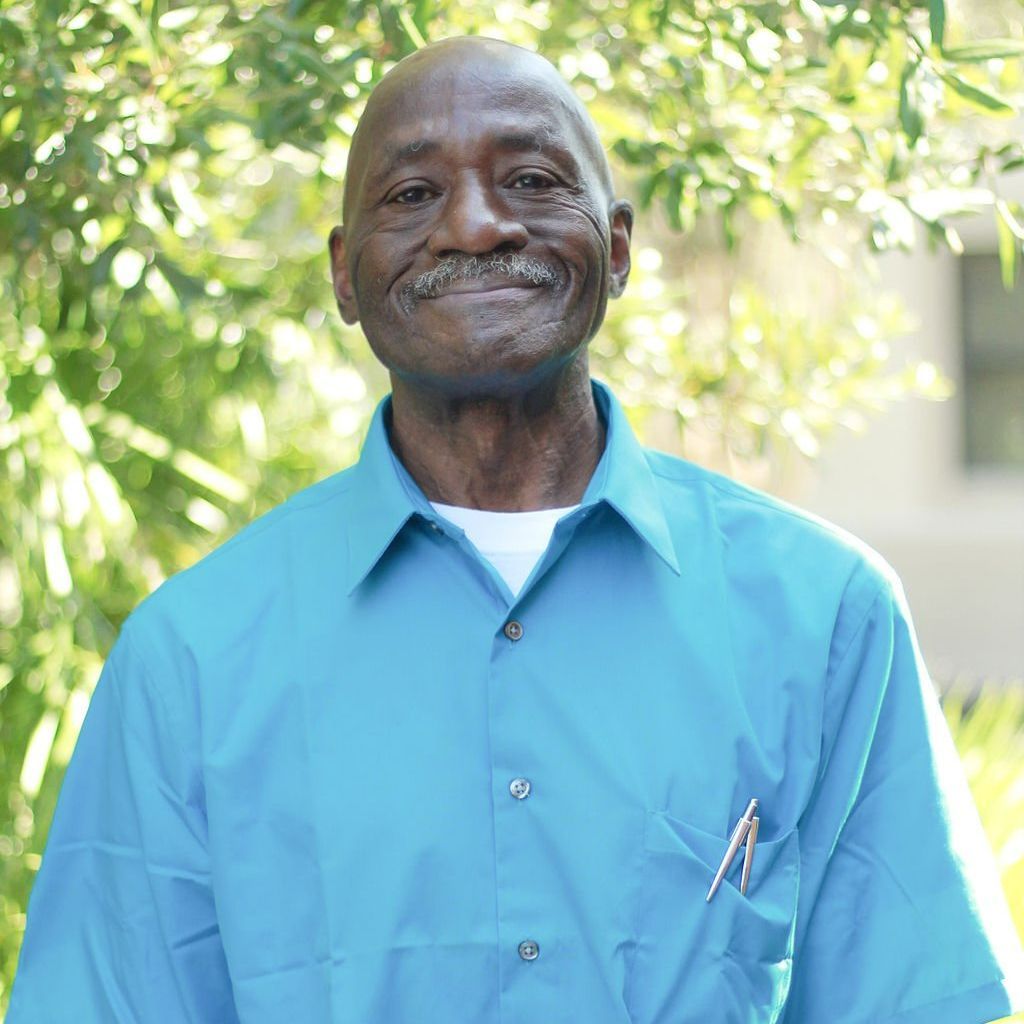 Smiling elderly Black man in a blue button-up shirt, outdoors in front of green foliage.