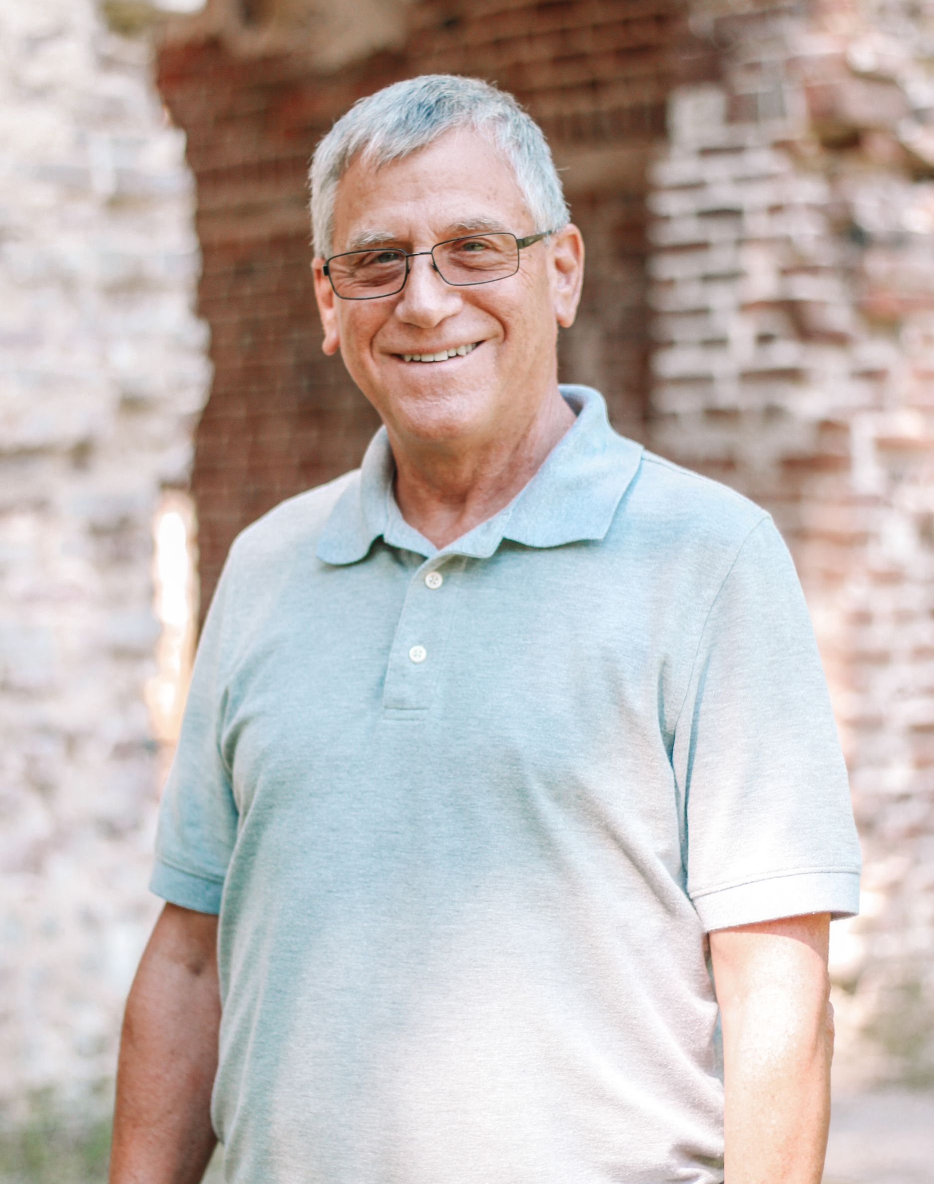 Man with gray hair and glasses smiles in front of stone ruins, wearing a light blue polo shirt.
