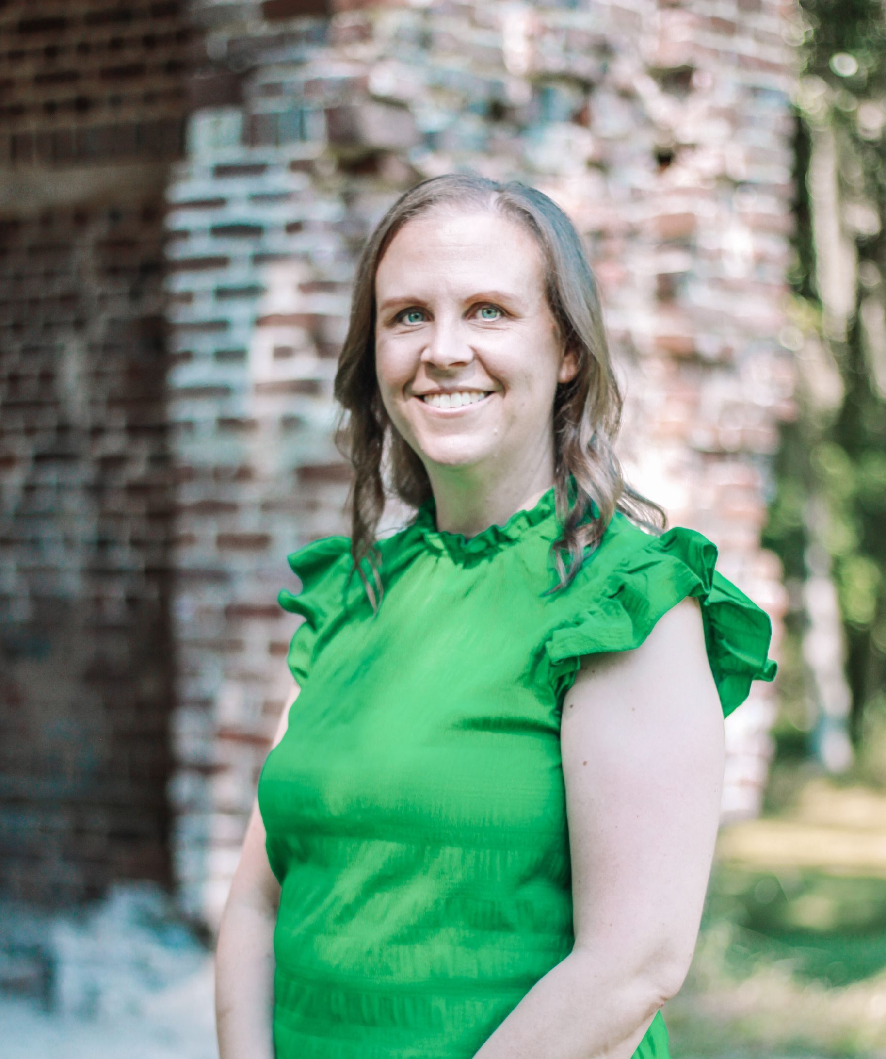 Woman with fair skin and light brown hair smiles in a green ruffled top, brick building in the background.