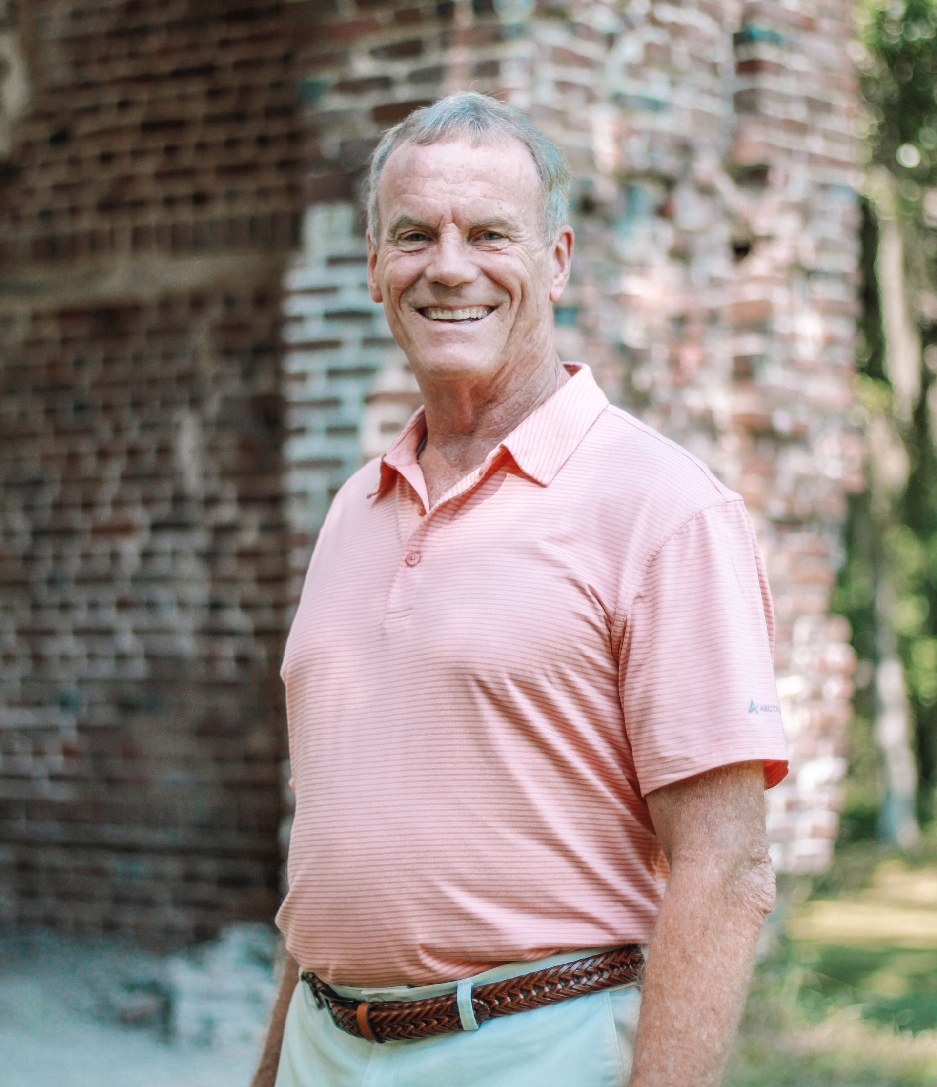 Man in a pink polo shirt smiles broadly, standing in front of weathered brick ruins.