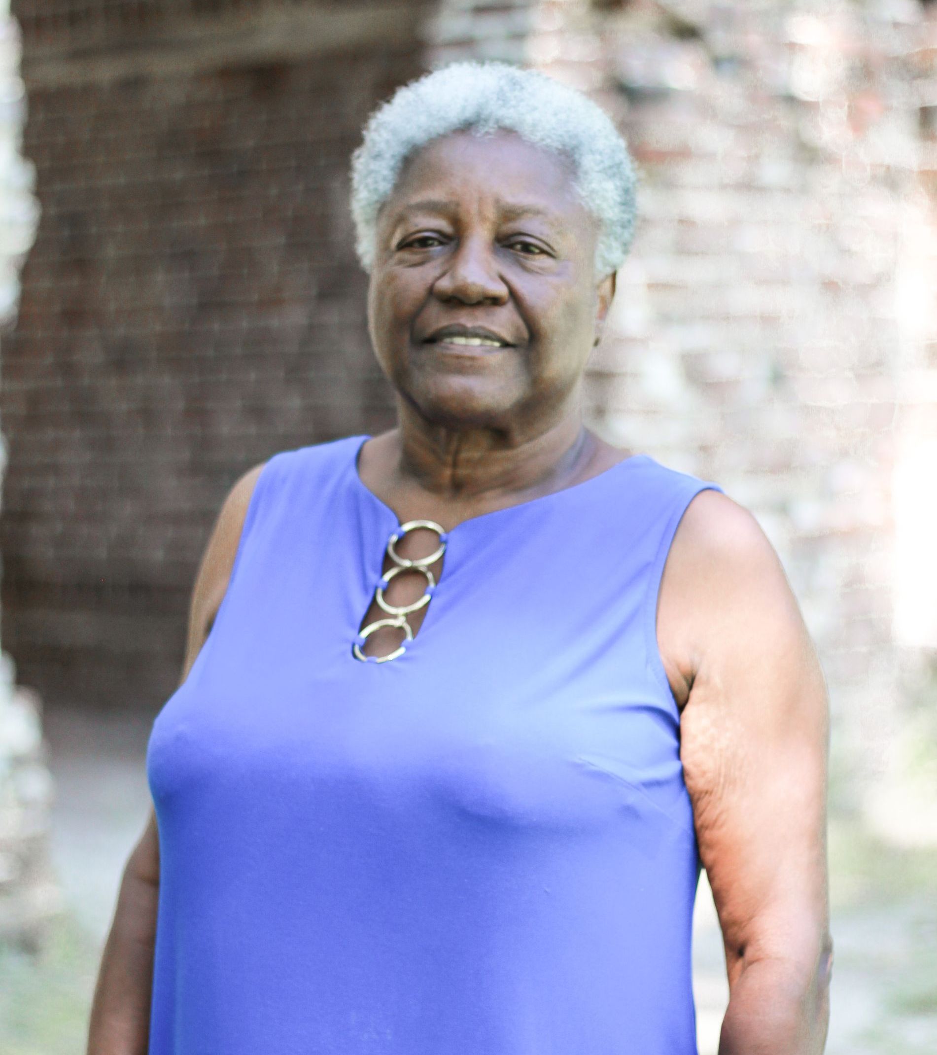 An older Black woman with short white hair smiles in a sleeveless blue dress, standing outside near a stone wall.