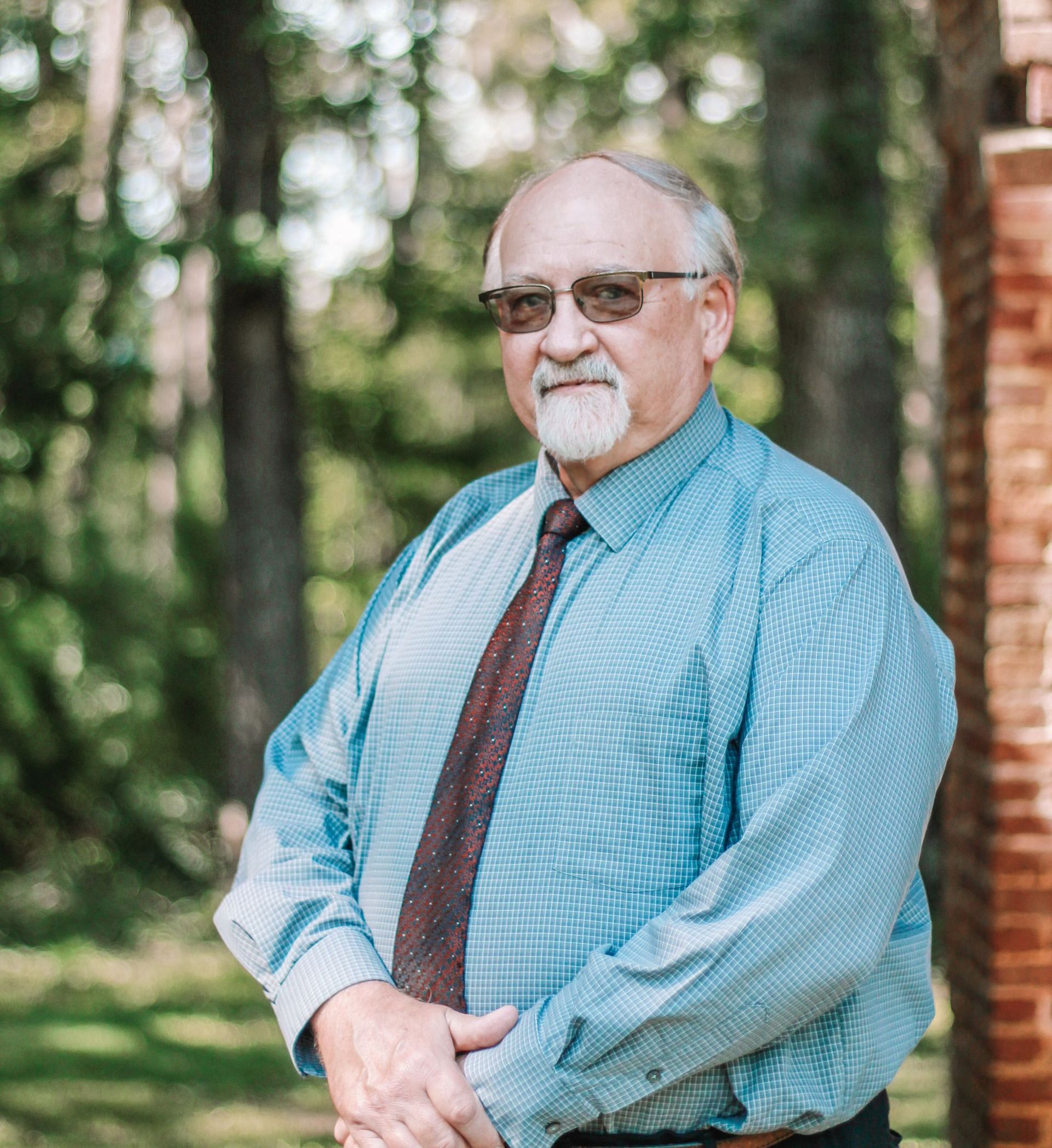 A man in a blue shirt and tie, with glasses and a goatee, stands outdoors with hands clasped, near a brick building.