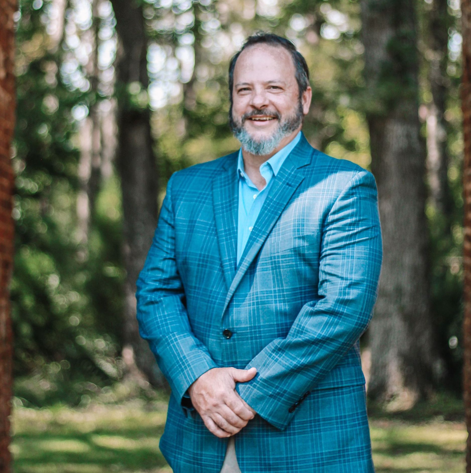 A man with a beard smiles, wearing a blue blazer and shirt, standing outdoors near trees.