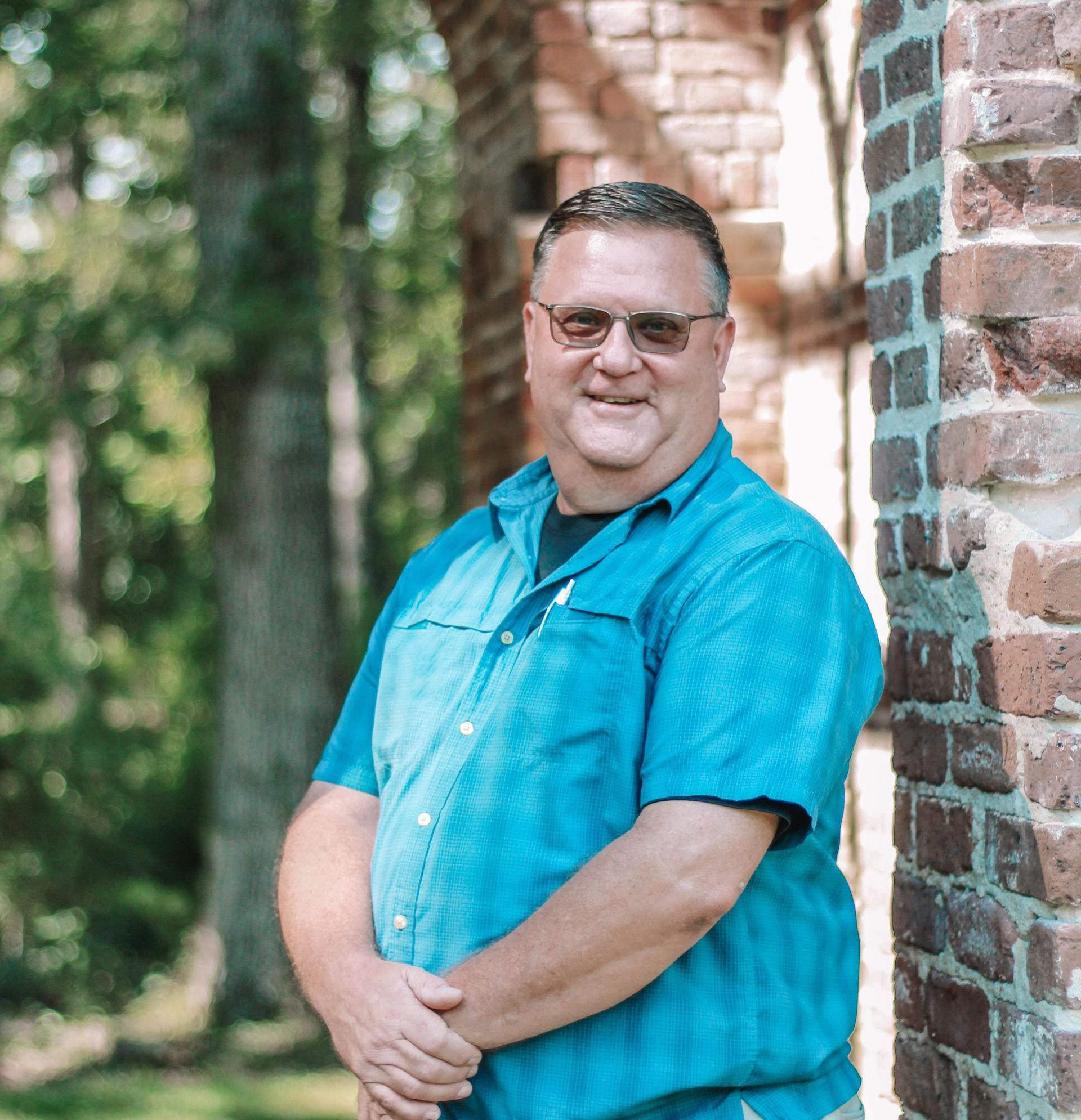 Man with glasses wearing a blue shirt, smiling, standing next to a brick wall in an outdoor setting.