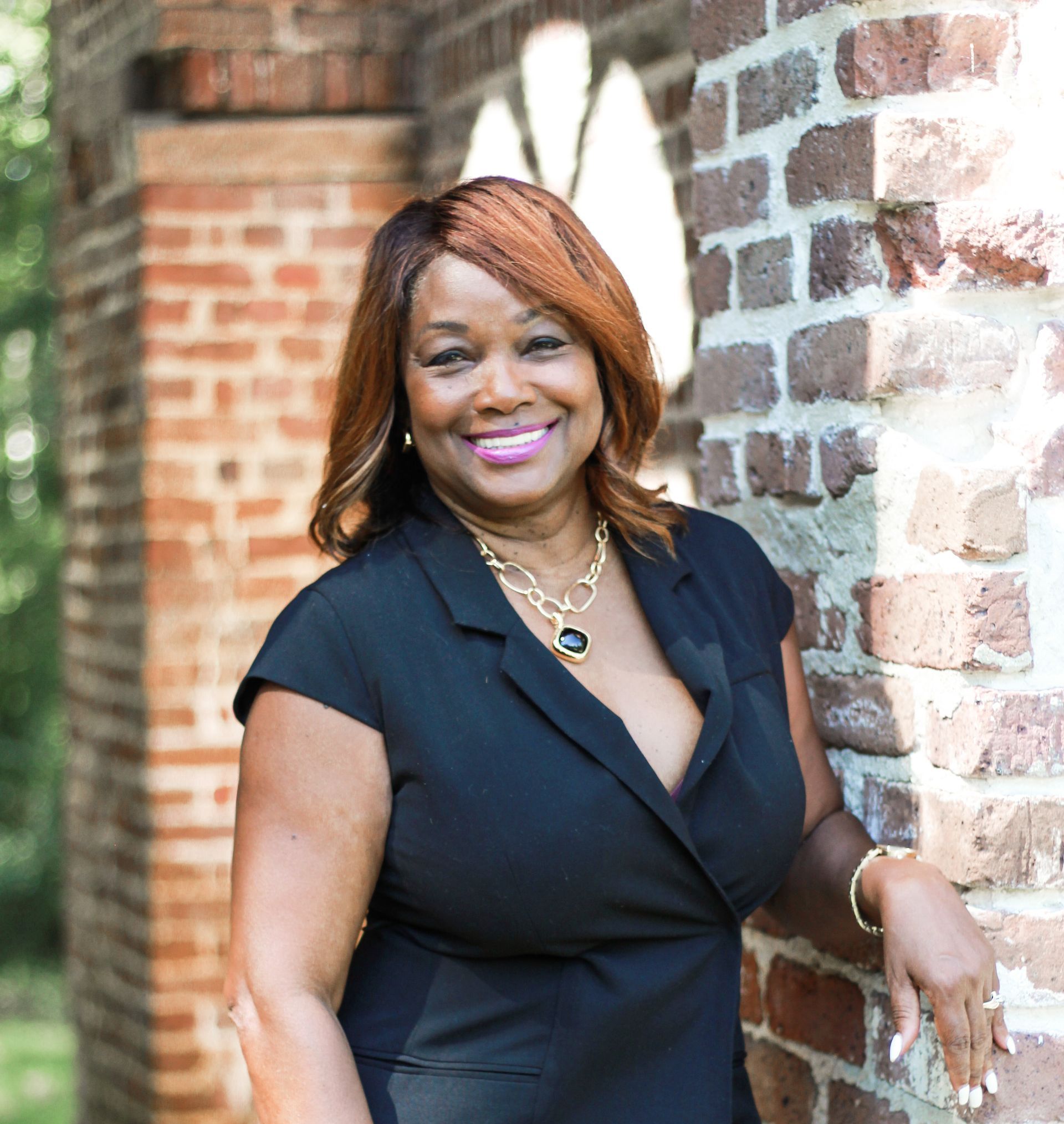 A smiling Black woman in a black dress leans against a brick wall. She has brown hair and a gold necklace.