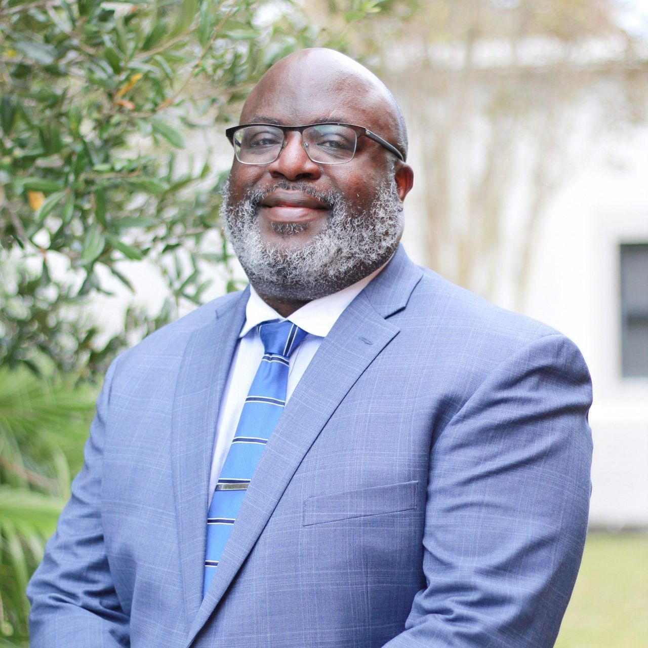 A Black man with a gray beard and glasses smiles, wearing a blue suit and tie, standing outside.