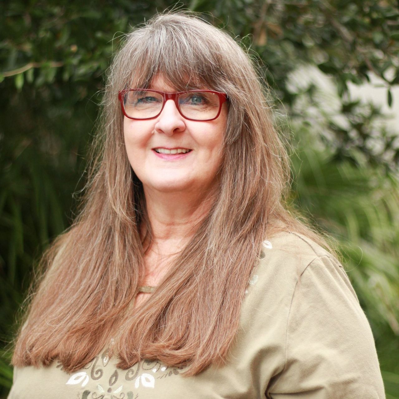 Woman with long brown hair and glasses smiles outdoors. She wears a light green shirt.