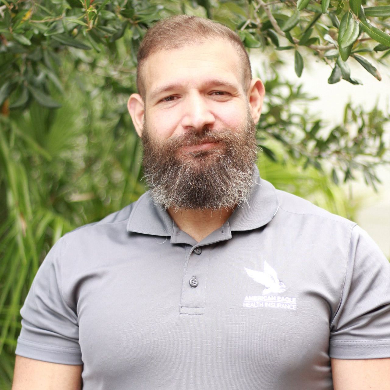 A man with a full beard wearing a gray polo shirt smiles slightly, standing outside with greenery in the background.