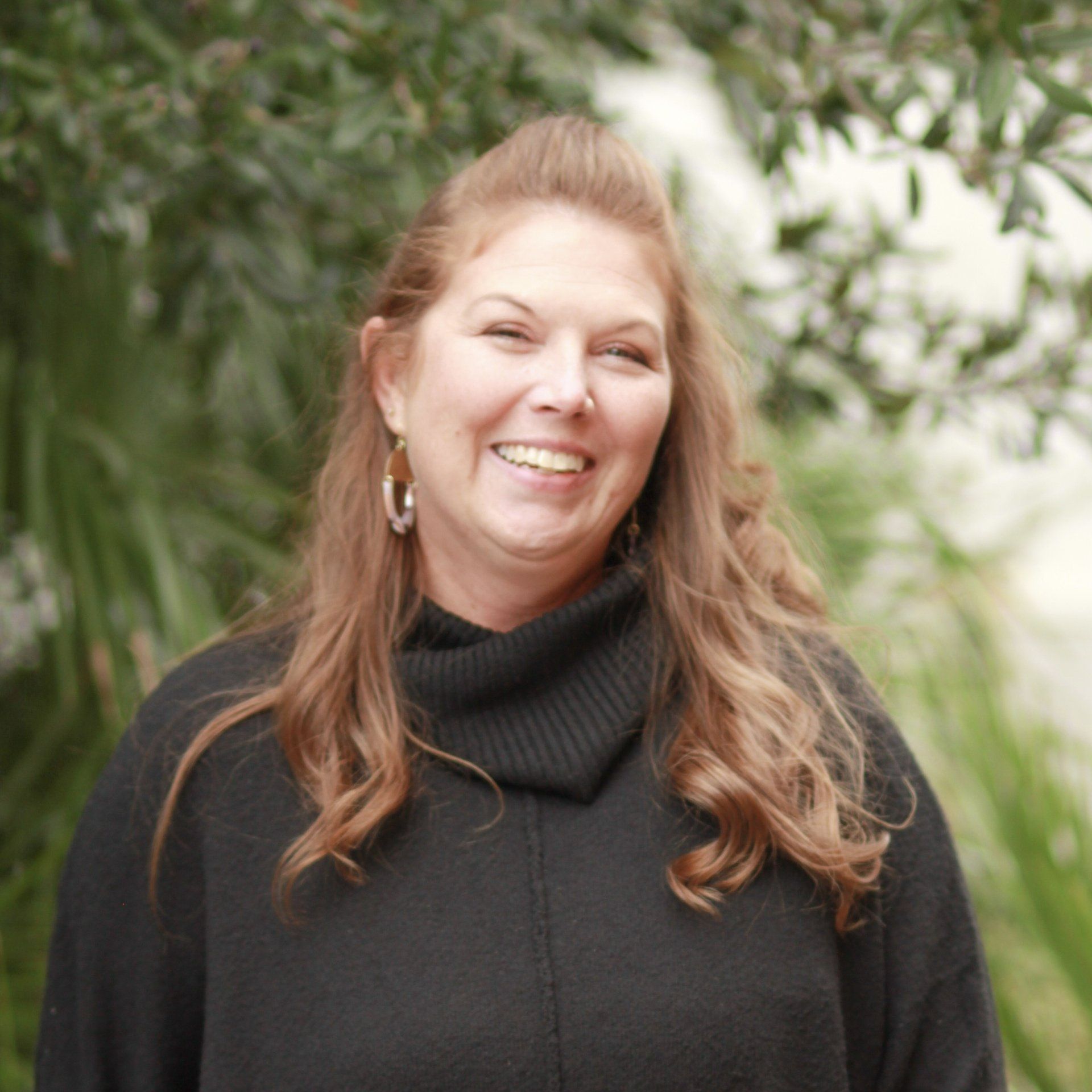 Woman with long wavy hair, smiling, wearing a black sweater, standing outside with foliage in the background.