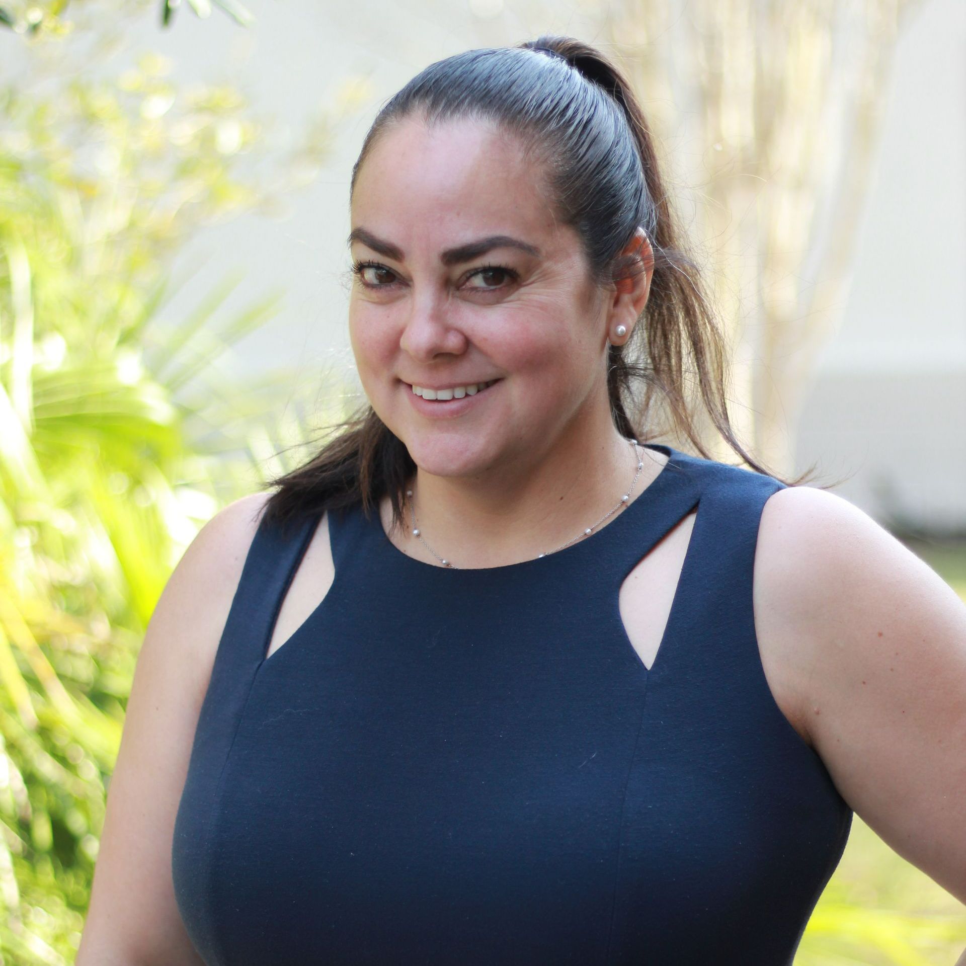 Woman with dark hair in a navy dress smiles outdoors. Green foliage and a white building are in the background.