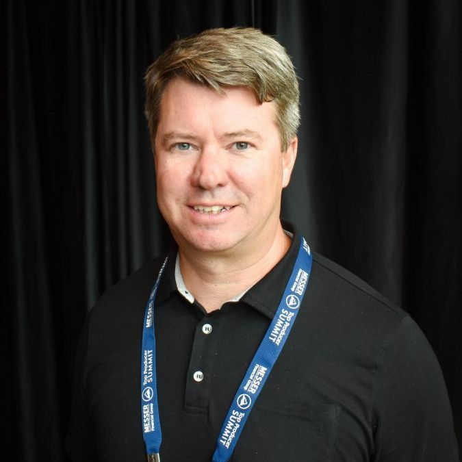 Man with fair skin and graying hair wearing a black polo shirt and lanyard, smiling against a dark curtain background.