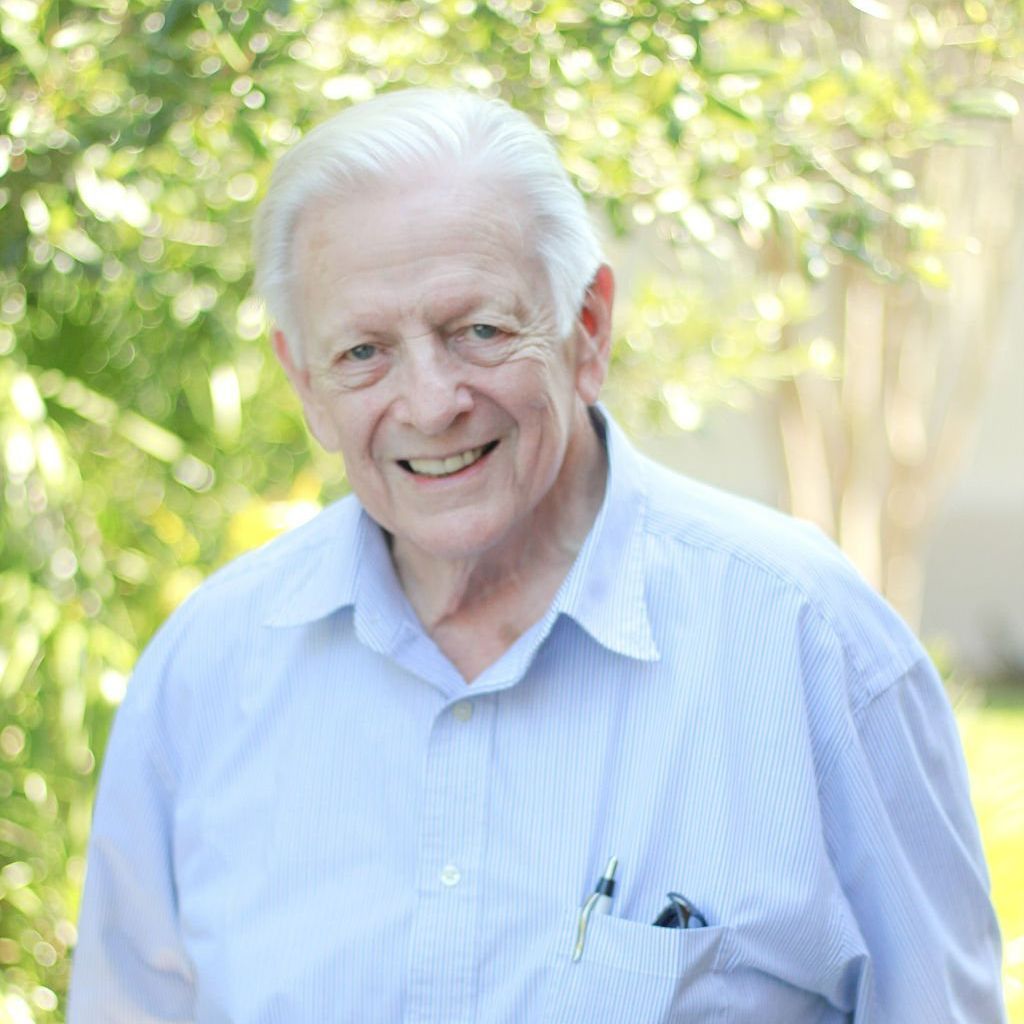 Smiling elderly man in a blue shirt, outdoors against a blurred green background.