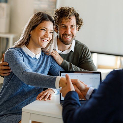 A smiling couple shaking hands with a person, possibly a financial advisor, at a table. The couple is in an office setting.