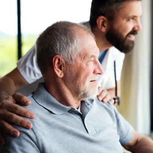 An older man in a gray shirt looks out a window while a younger man with a beard and stethoscope puts his hand on the older man's shoulder.