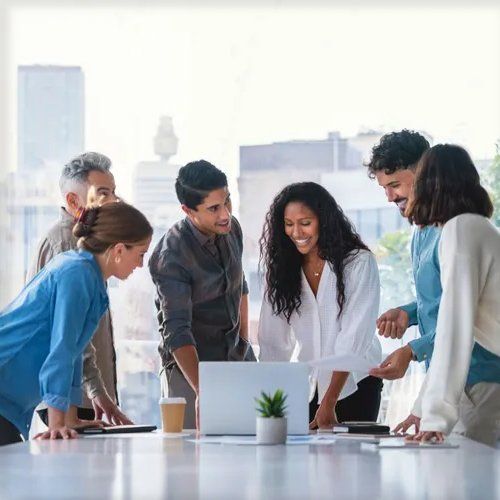Group of diverse colleagues collaborating around a table in a bright office, looking at a laptop and documents with smiles.