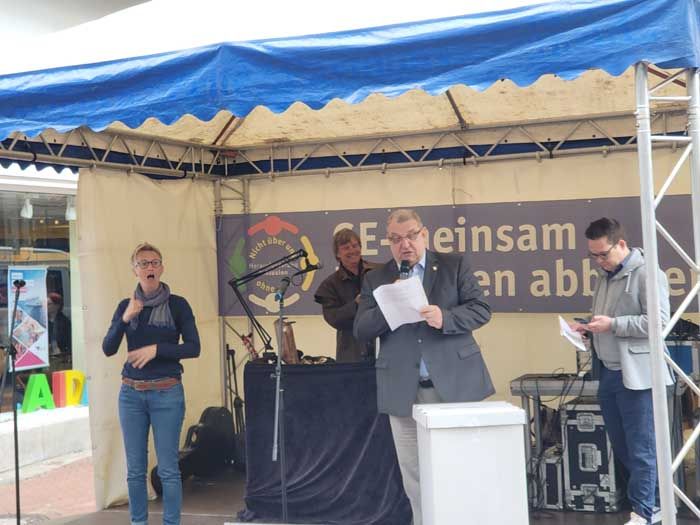 Unter einem Pavillon befindet sich ein Messestand der AGB Gelsenkirchen. In der Mitte steht der Vorsitzende Dieter Harwardt mit einem Mikrofon und einem Redemanuskript. Links von ihm steht eine Gebärdendolmetscherin.