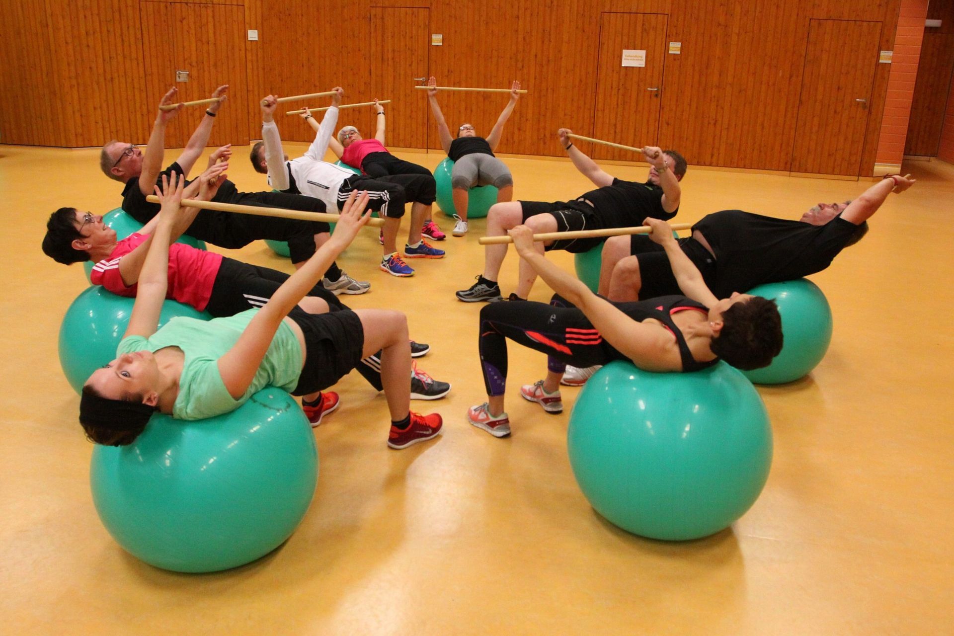 Menschen betreiben Gymnastik. Sie liegen mit dem Rücken auf großen Sitzbällen und Recken die Arme in die Höhe. Sie halten dabei jweiles eine Holzstange. Die Szene spielt sich in einer Turnhalle ab,