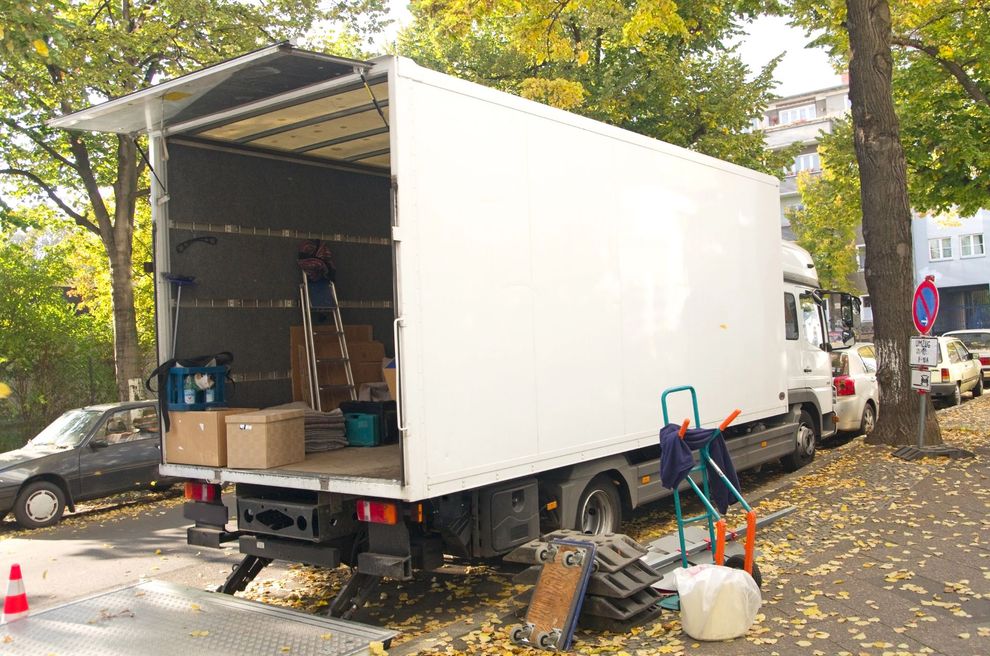 Un furgone bianco con cassone aperto è parcheggiato su una strada alberata, con scatole e materiale per traslochi a terra.