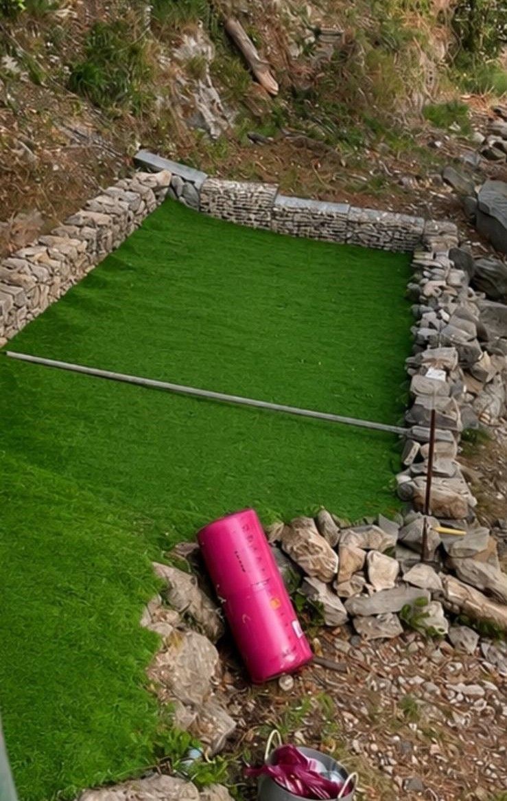 Piccolo putting green in erba sintetica, delimitato da rocce, con un cilindro rosa appoggiato nelle vicinanze.
