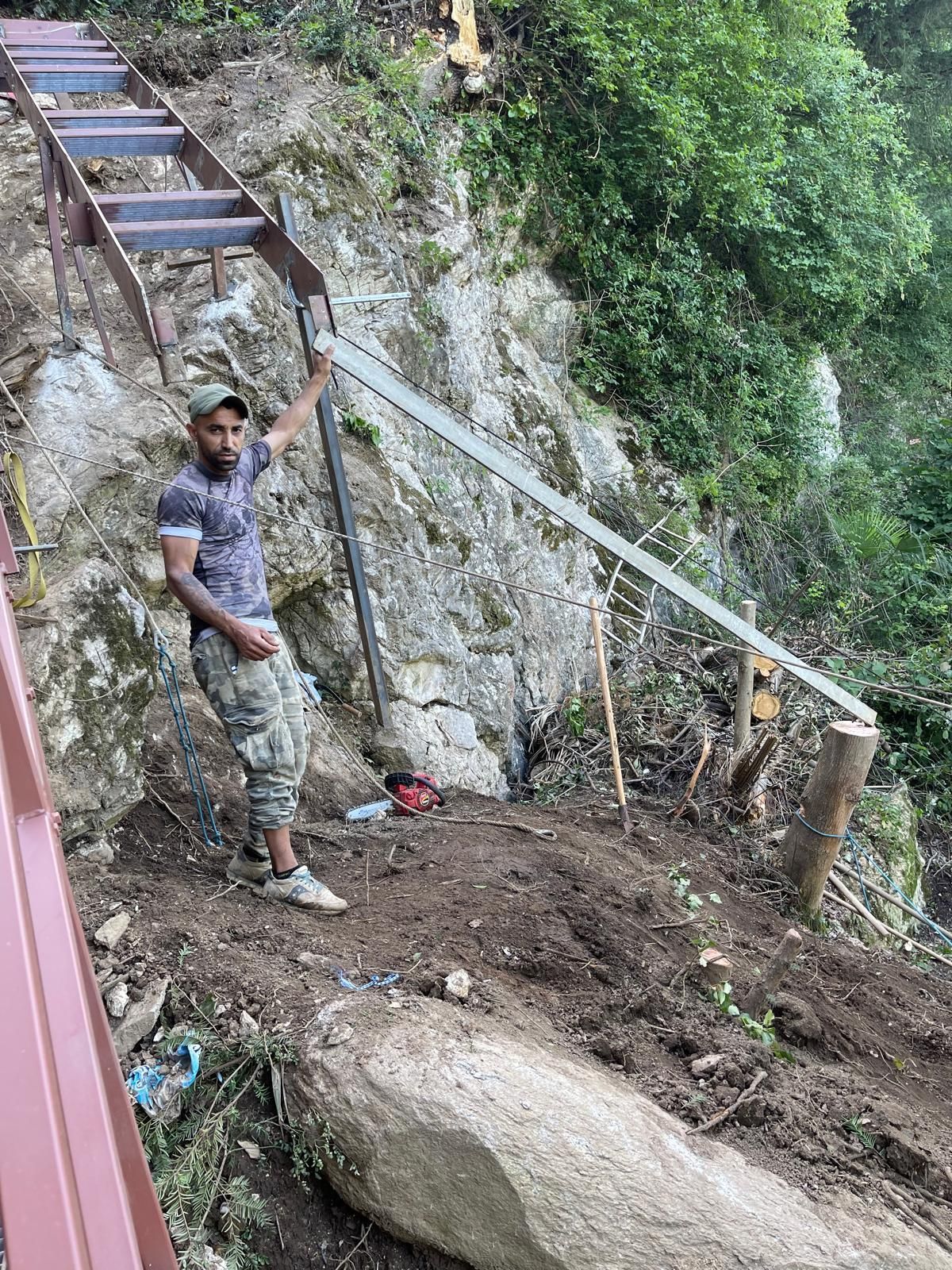 Un uomo in piedi lungo un ripido sentiero roccioso con scale e ringhiera in metallo, in una zona boscosa.